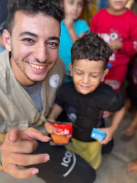 Mohammed wearing a Gaza Food Relief Project beige vest with a grey vest underneath amd black pants. He is smiling at the camera while holding a small child. The child is wearing a black shirt and yellow pants. He hass his eyes closed and is holding a snack that Mohammed fundraised for. Two children are in the background; one with a blue outfit and one with a red outfit.