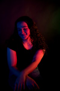 a woman with long curly hair sitting down in front of a colorful background
