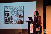 a woman standing at a podium with a drawing on the screen