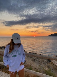 a woman wearing a hat and shorts standing next to the ocean at sunset