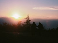 two people standing on top of a hill with the sun setting behind them