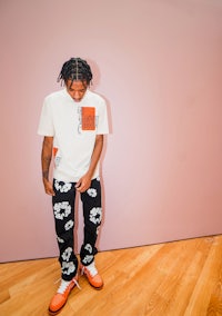a man standing in front of a pink wall wearing a t - shirt and floral pants