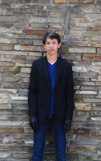 a young man in a black jacket leaning against a brick wall
