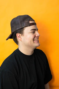 a young man wearing a black hat and smiling against an orange wall