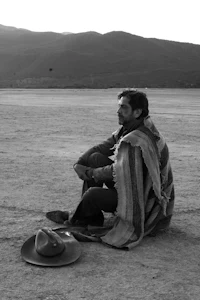 a man sitting in the desert with a blanket and hat