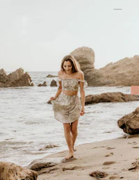 a young woman walking on the beach with rocks in the background