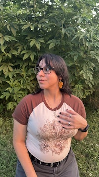 a woman wearing glasses and a t - shirt posing in front of bushes
