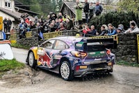 a rally car driving down a dirt road in front of a crowd