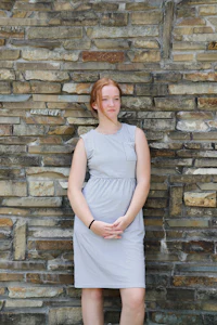 a woman in a gray dress leaning against a stone wall