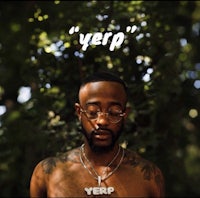 a man with tattoos and glasses standing in front of trees