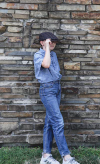a boy in jeans and a hat leaning against a stone wall