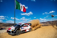 a rally car driving down a dirt road with a helicopter flying over it