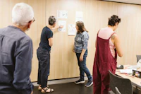 Four adults participate in a creative workshop. A woman in a navy T-shirt points to a drawing displayed on a wood-panelled wall while another woman examines the artwork. An older man observes from the foreground, and a woman in burgundy dungarees stands beside a table covered with art materials. Several sketches and colourful drawings are displayed on the wall. Image courtesy of Alexandra Szigeti