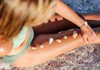 a woman in a bikini sitting on the sand with seashells on her legs
