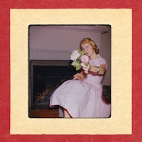 a woman in a pink dress holding flowers in front of a fireplace