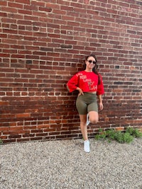 a woman leaning against a brick wall in a red sweater and shorts