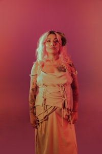 a woman in a white dress with tattoos standing in front of a pink background