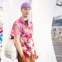 a man wearing a floral shirt and purple hat standing next to a tote bag