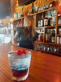 a woman standing in front of a bar with a drink in front of her