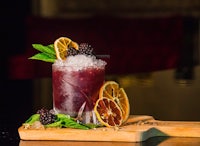 a cocktail with blackberries and mint on a wooden cutting board