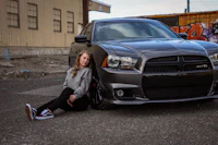 a girl sitting on the ground next to a gray dodge charger