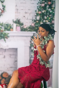 a woman in a red dress drinking wine in front of a christmas tree