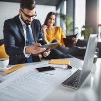 An image featuring two people, one man in a business suit, the other a woman and a very professional outfit. They are sitting in an office environment, looking over paperwork and looking at information on the tablet.