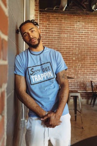 a man leaning against a brick wall wearing a blue t - shirt