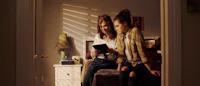two women sitting in a room looking at a tablet