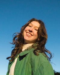 Grace, a young white woman, is smiling towards the camera. She is wearing a cream turtleneck with a bright green jacket and her hair is down. The blue sky is behind her.