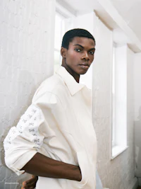 a black man in a white jacket leaning against a wall