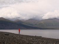 a man is standing on the shore of a lake with mountains in the background