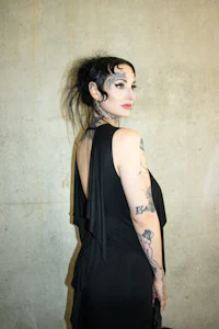a woman in a black dress with tattoos posing in front of a wall