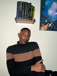 a man in a sweater sitting in front of a book shelf
