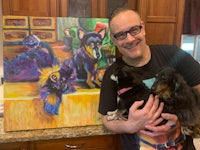 a man holding a painting of two dogs in front of a kitchen