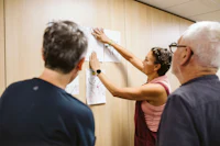 A woman in a pink top and burgundy dungarees gestures towards several drawings displayed on a wood-panelled wall, while two men watch and listen. The three appear engaged in a discussion about the artwork in a brightly lit creative workshop space. Image courtesy of Alexandra Szigeti
