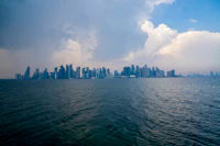 A panoramic view of Doha’s modern skyline from across the Arabian Gulf, with towering skyscrapers set against a cloudy sky.