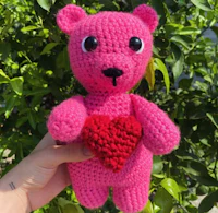 a pink crocheted teddy bear holding a red heart
