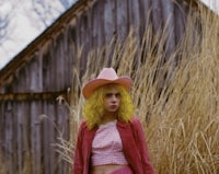 a girl in a pink dress and hat standing in front of a barn