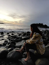 a woman sitting on rocks near the ocean at sunset