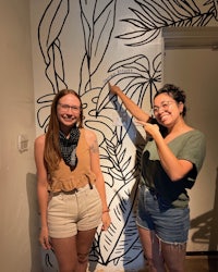 two women standing in front of a wall with a black and white drawing
