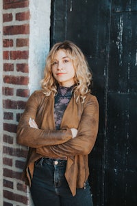 a woman in a tan jacket leaning against a brick wall