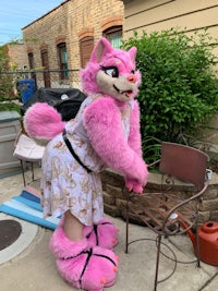 a pink mascot dressed in a dress standing next to a chair