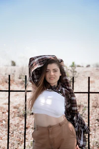 a woman in a plaid shirt and tan pants leaning against a fence