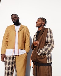 two men standing next to each other wearing tan pants and jackets