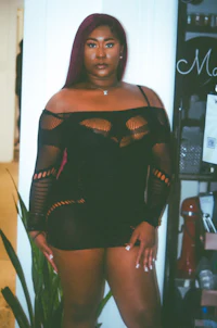 a black woman in a mesh dress posing in front of a plant