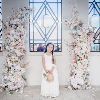 a little girl in a white dress standing in front of a floral arch