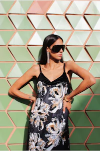 a woman wearing sunglasses and a floral dress leaning against a wall