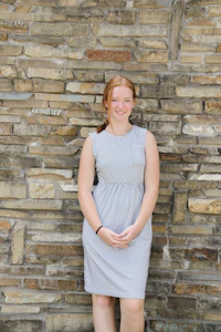 a young woman in a gray dress leaning against a stone wall