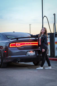 a woman standing next to a gray dodge charger at a gas station
